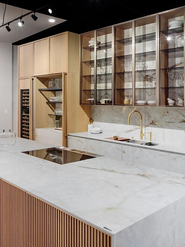 Modern Boston kitchen with light wood cabinetry, glass display shelves, marble countertops, and a gold faucet for a refined, elegant look.