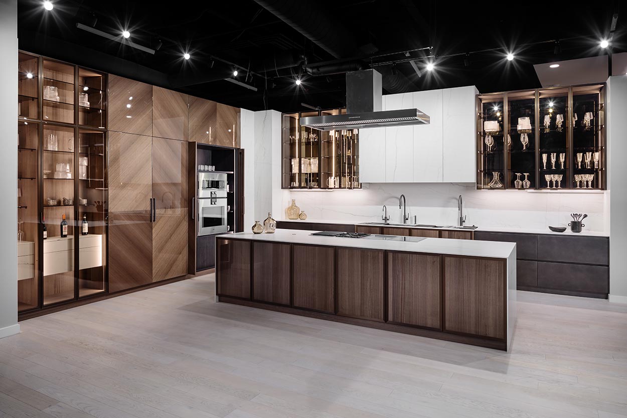 A modern luxury kitchen with wood cabinets, stainless steel appliances, white countertops, and elegant glass display cases illuminated by stylish ceiling spotlights.