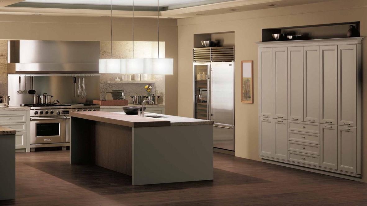 A well-lit kitchen with taupe cabinetry, stainless steel appliances, an island with a sink, and pendant lights, all set against a beige stone backsplash.