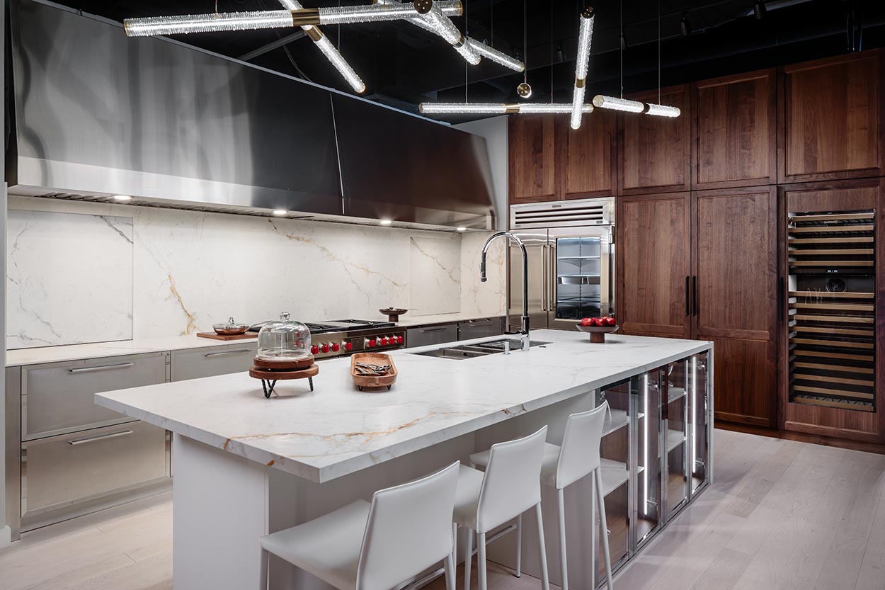 A modern kitchen featuring dark wood cabinetry, stainless steel appliances, white marble countertops, an island with seating, and unique linear pendant lights.