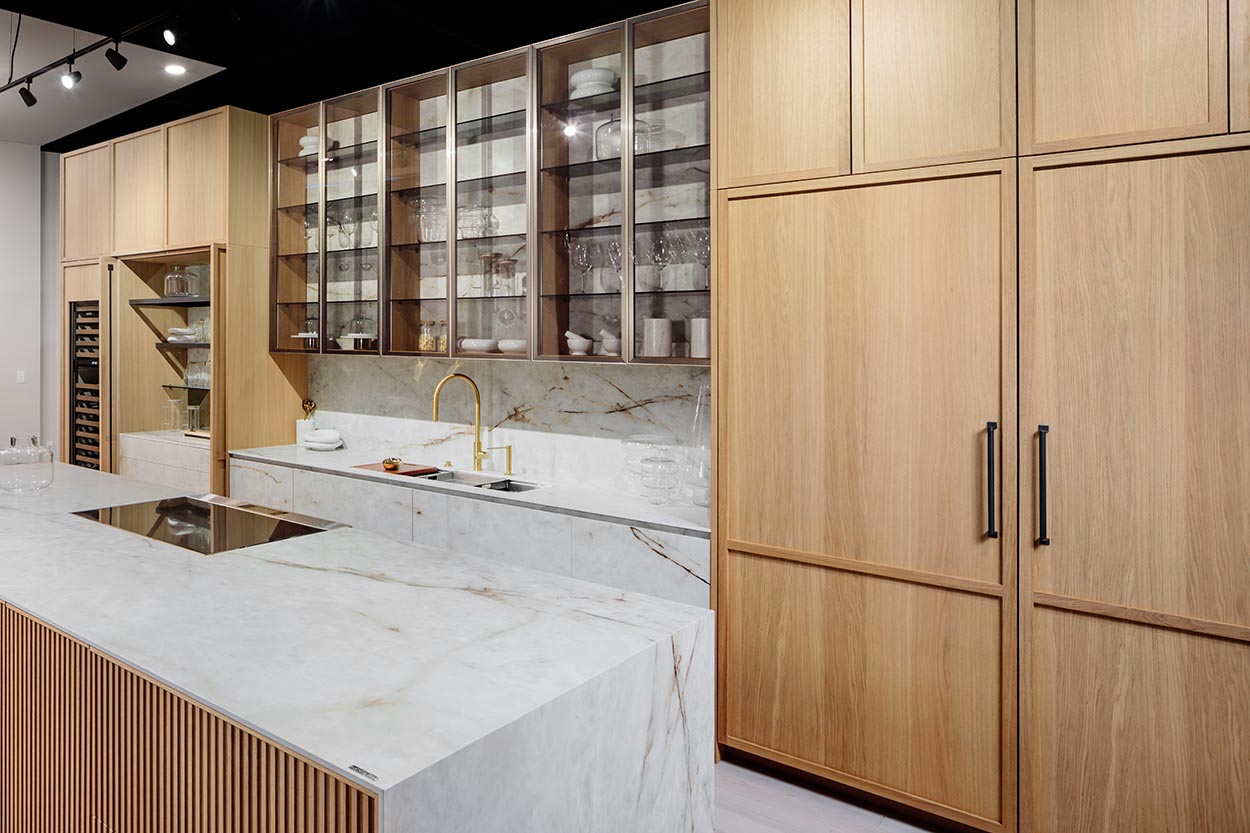 Modern Boston kitchen with light wood cabinetry, glass display shelves, marble countertops, and a gold faucet for a refined, elegant look.